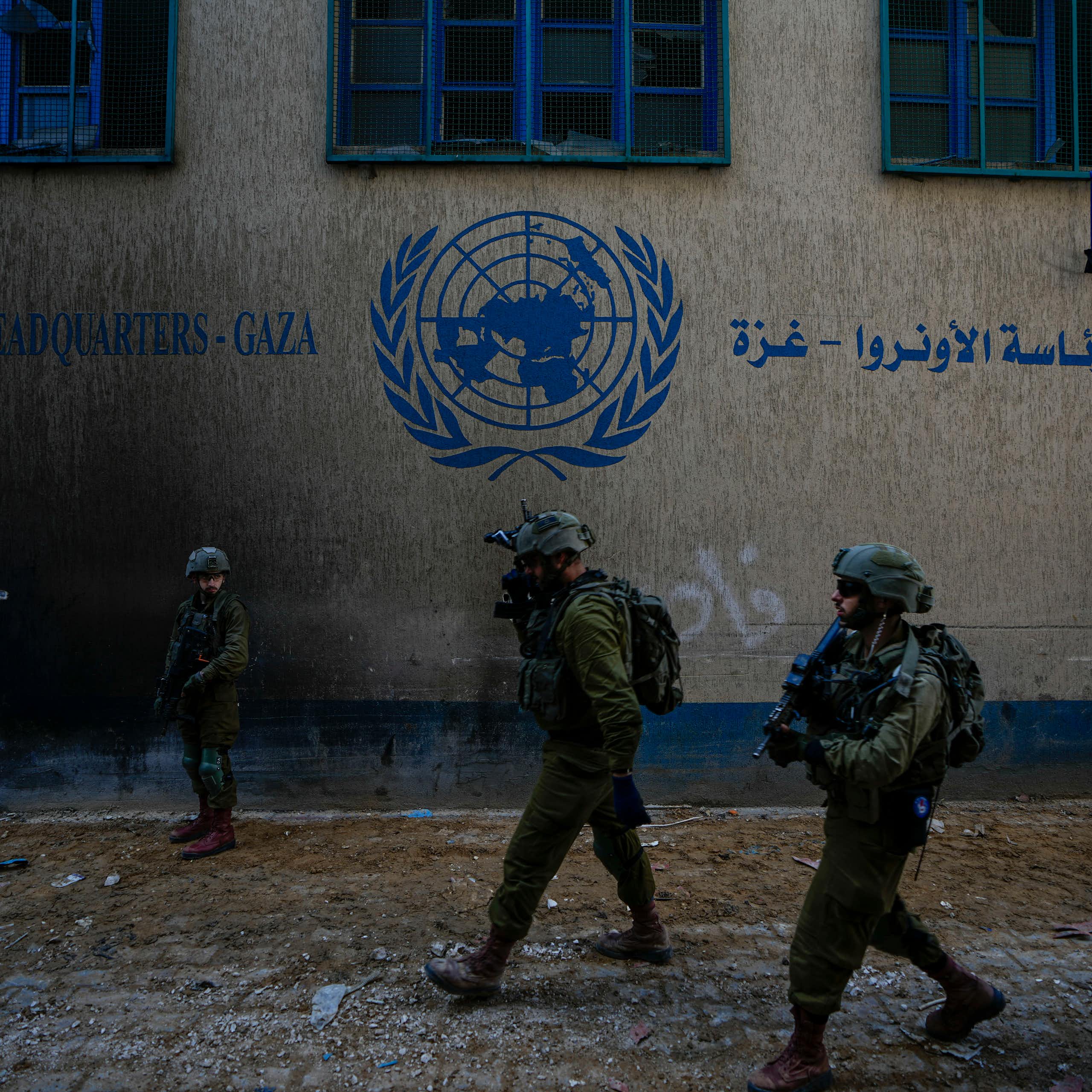Soldiers in fatigues and helmets walk past a building with a UN logo on it.