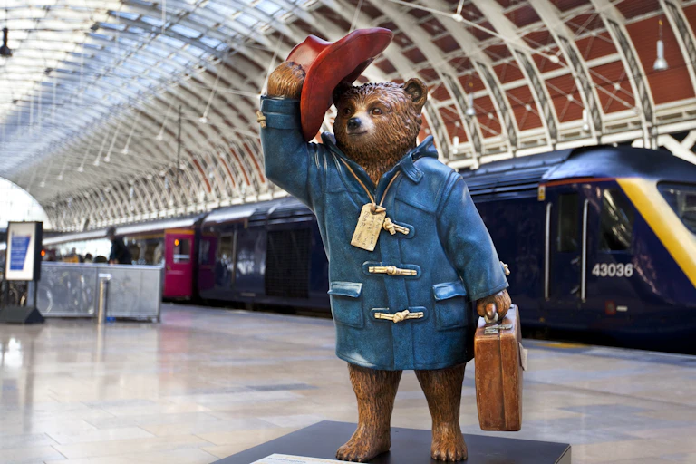 A statue of Paddington Bear carrying a suitcase and tipping his hat, on a platform at Paddington station