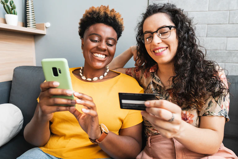 A young couple sitting on a couch looking at a bank card and phone, smiling