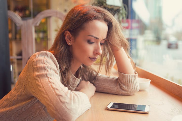 A young woman sitting at a cafe window, looking sadly at a mobile phone on the counter in front of her