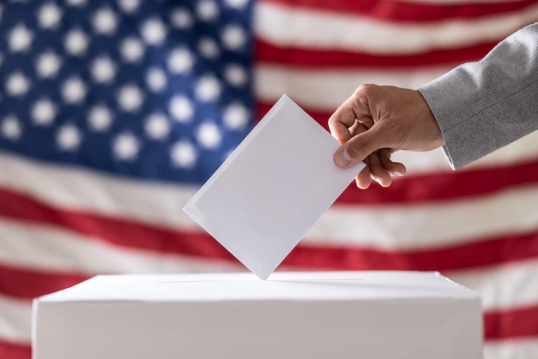 A person places a piece of paper in a box in front of a US flag.