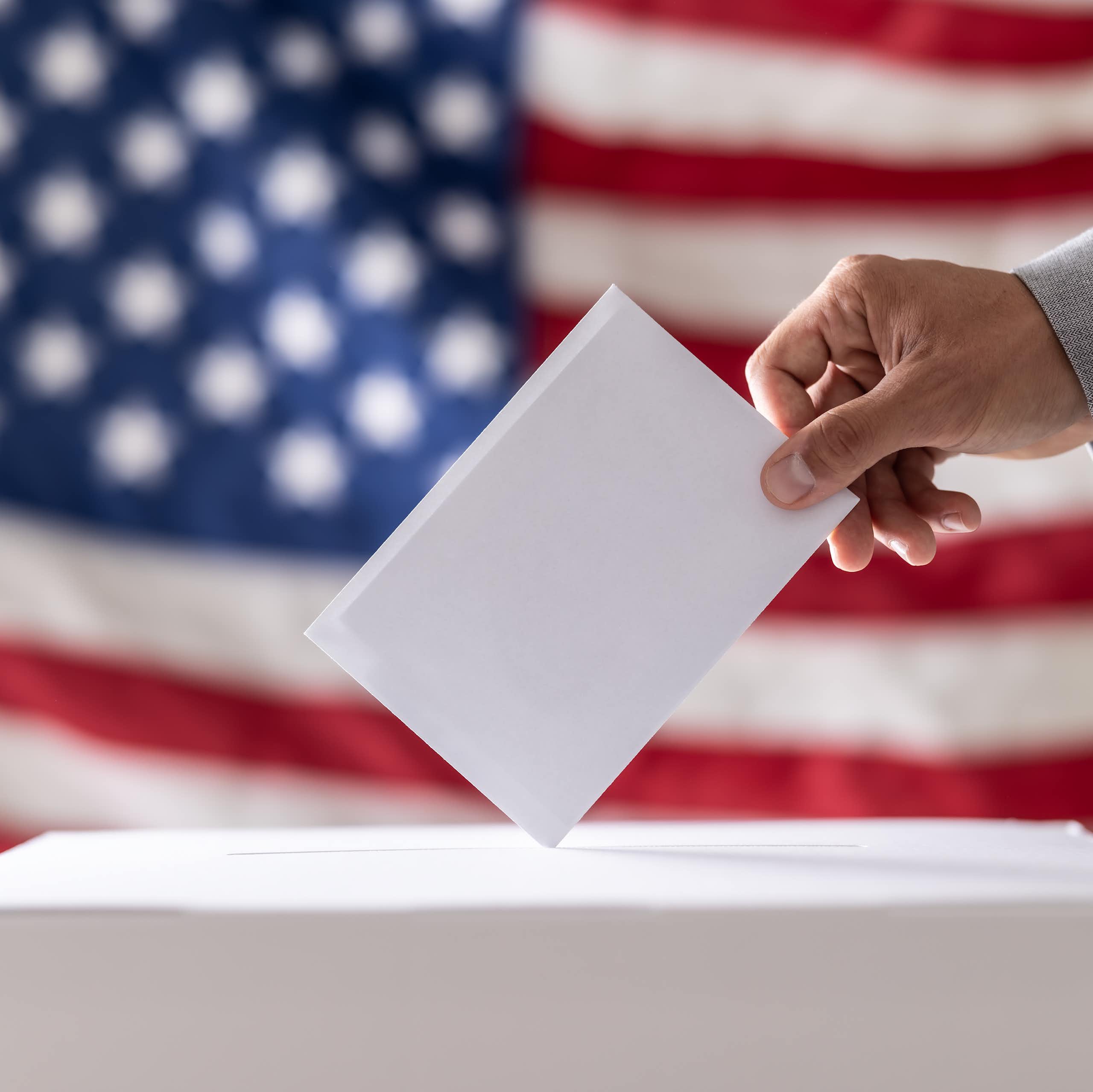 A person places a piece of paper in a box in front of a US flag.