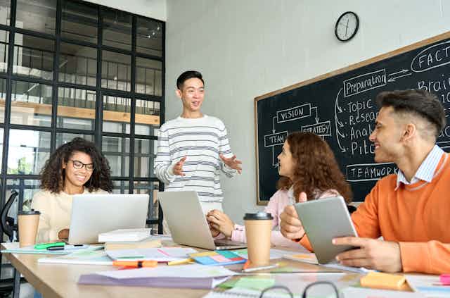 A young man stands while speaking to a group of peers who are sitting around a table