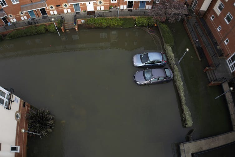 Cars sit stranded in a flooded car park surrounded by buildings.