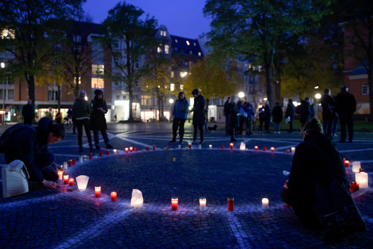 People in dark coats stand around a circle of lit candles on a cobblestone plaza with a geometric design built into it as dusk falls.