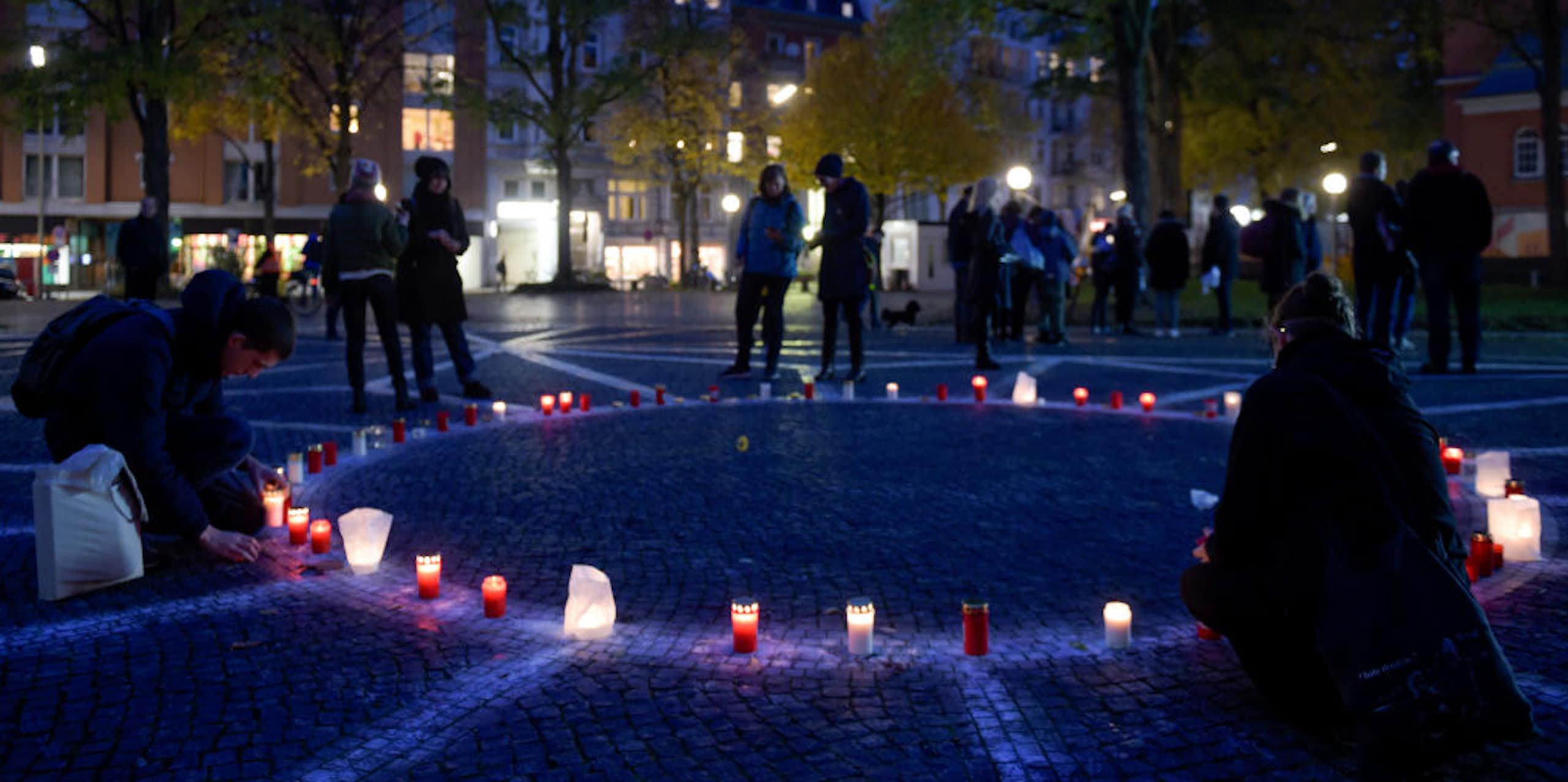 People in dark coats stand around a circle of lit candles on a cobblestone plaza with a geometric design built into it as dusk falls.