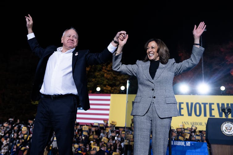 A white man in a black jacket and white shirt holds up the hand of a brown skinned woman with a grey pantsuit in front of a large crowd of people. Behind them is an American flag and a yellow banner that says 'Vote'