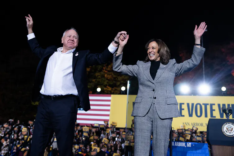 A white man in a black jacket and white shirt holds up the hand of a brown skinned woman with a grey pantsuit in front of a large crowd of people. Behind them is an American flag and a yellow banner that says 'Vote'