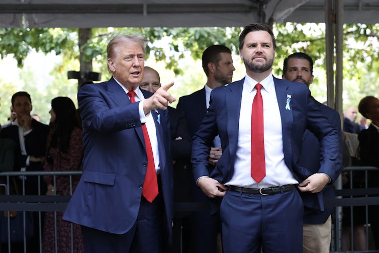 Two white men wear navy blue suits, white shirts and red ties and look ahead.