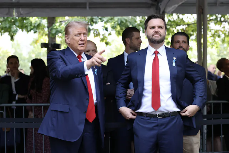 Two white men wear navy blue suits, white shirts and red ties and look ahead.