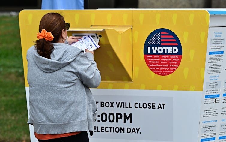 A woman with a brown ponytail is seen from behind standing in front of a large yellow and white box that says 'I voted.'