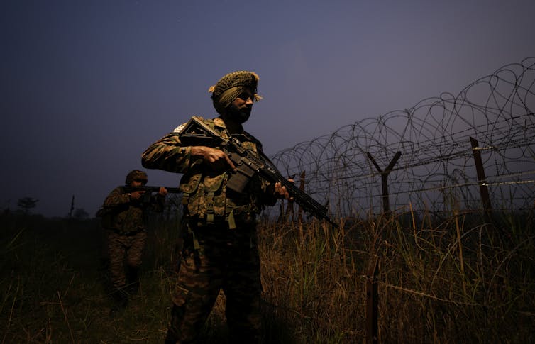 A soldier wearing a turban and in camouflage carrries a rifle and stands next to a barbed wire fence.
