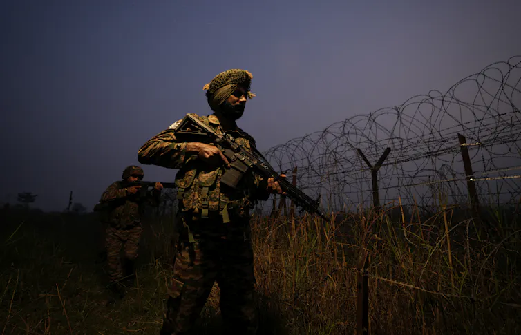 A soldier wearing a turban and in camouflage carrries a rifle and stands next to a barbed wire fence.