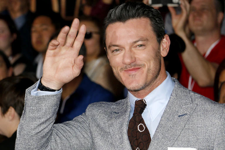 Hollywood actor Luke Evans smiles and waves at camera. He's wearing a grey suit with a brown patterned tie and is standing in front of a crowd of people.