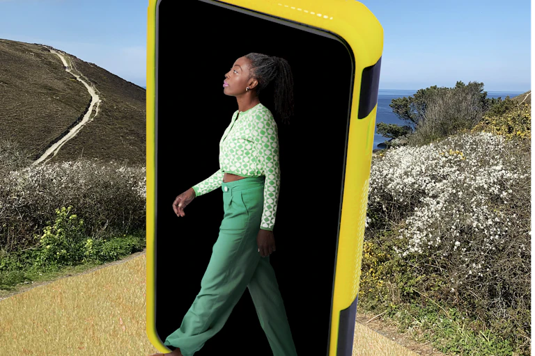 a woman appears to emerge from a giant smartphone onto a path in a nature preserve