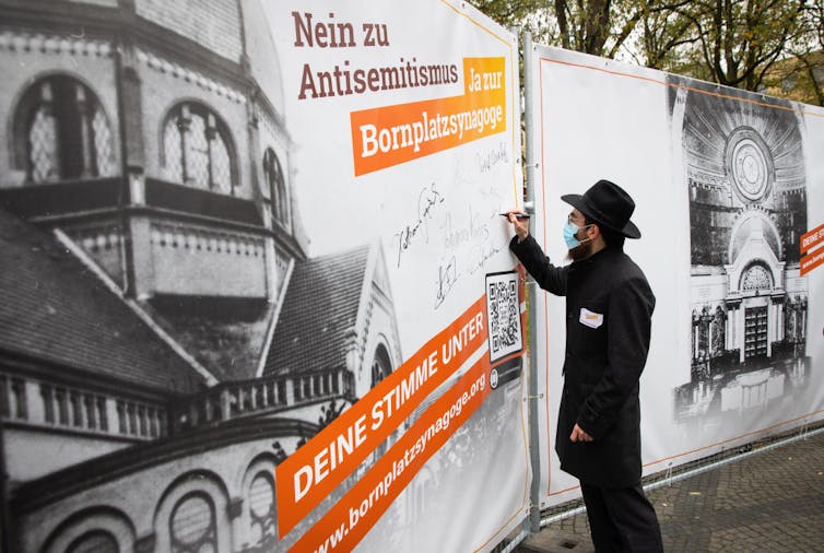 A man in a black outfit and black hat signs a large poster with a photo of a building and orange text.