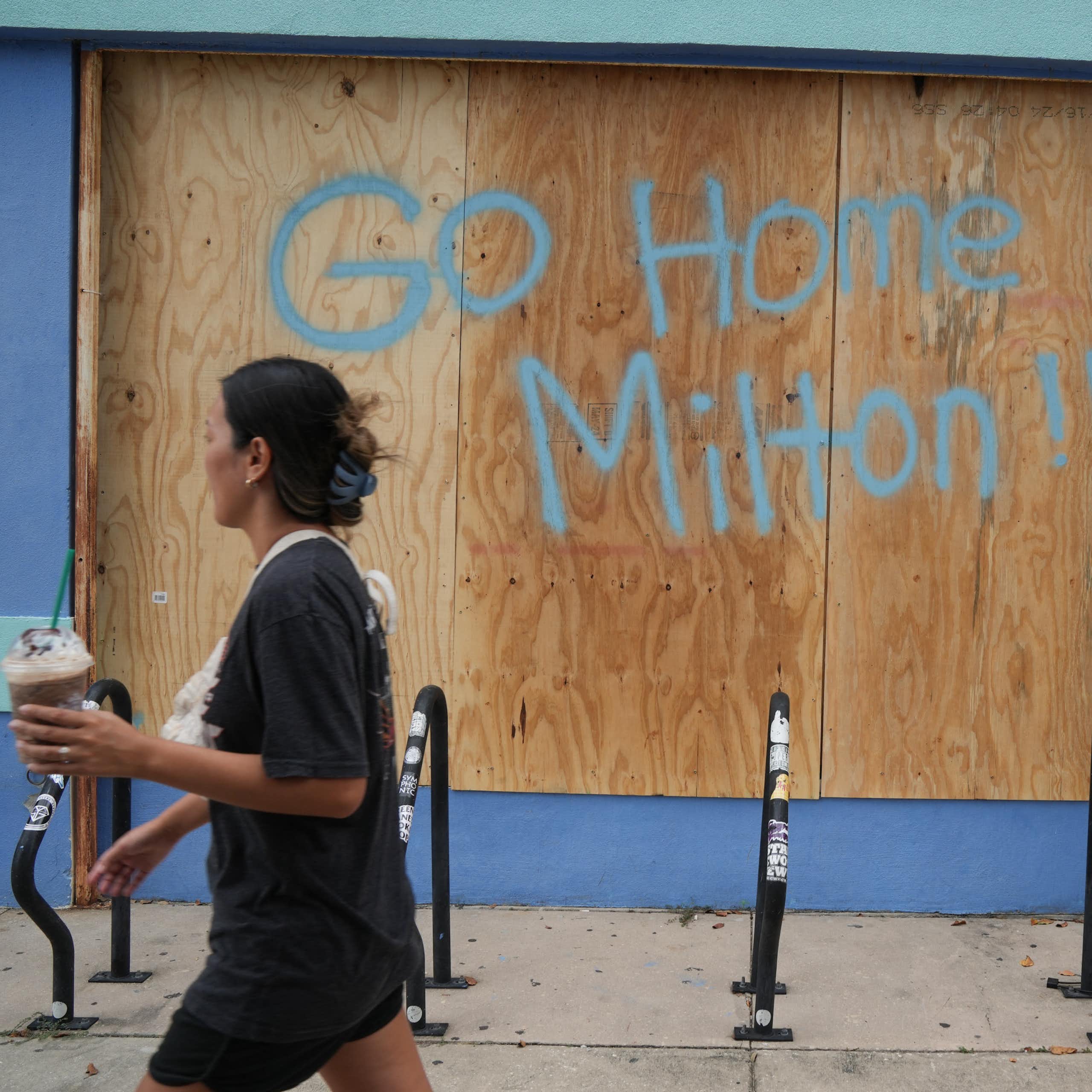 People carrying coffee drinks walk by a sign scrawled on plywood-covered windows reading "Go home Milton!"