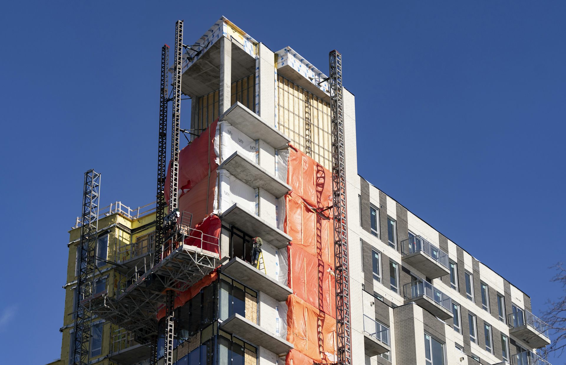 A man stands on a ladder on a platform suspended on the side of a condo building, which is under construction