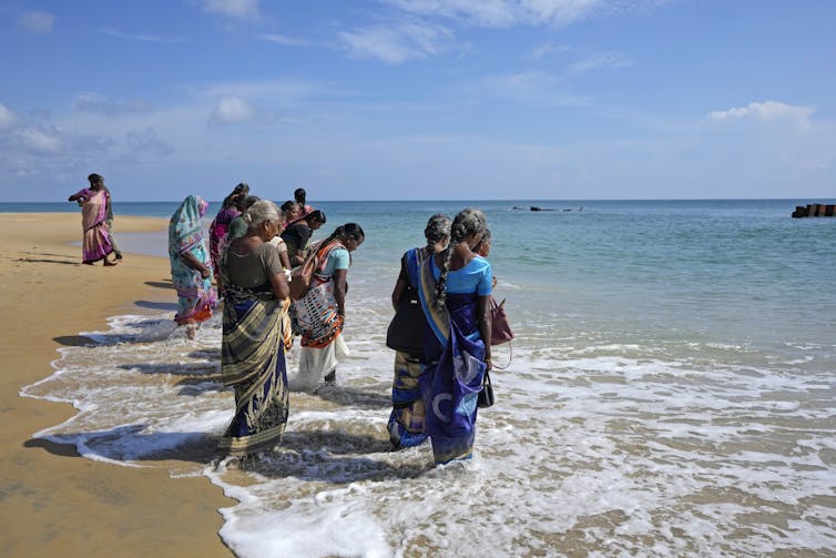 Women in bright garb stand on the shore of a beach.