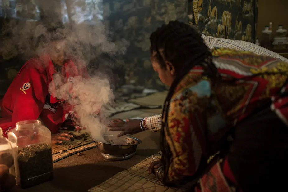 Two people, one wearing beaded bracelets up their arm, sit next to a silver bowl from which smoke is pouring as they perform a ritual