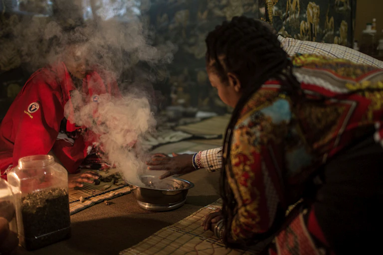 Two people, one wearing beaded bracelets up their arm, sit next to a silver bowl from which smoke is pouring as they perform a ritual