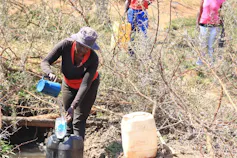 A woman uses a jug to draw water from a hand dug well in the soil, and pours water through a funnel into a 20 litre container