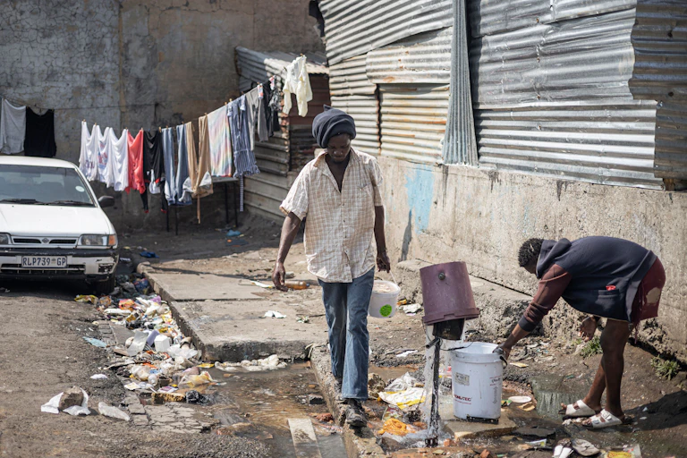 A broken tap with water spurting in different directions and a woman trying to collect the water with a bucket