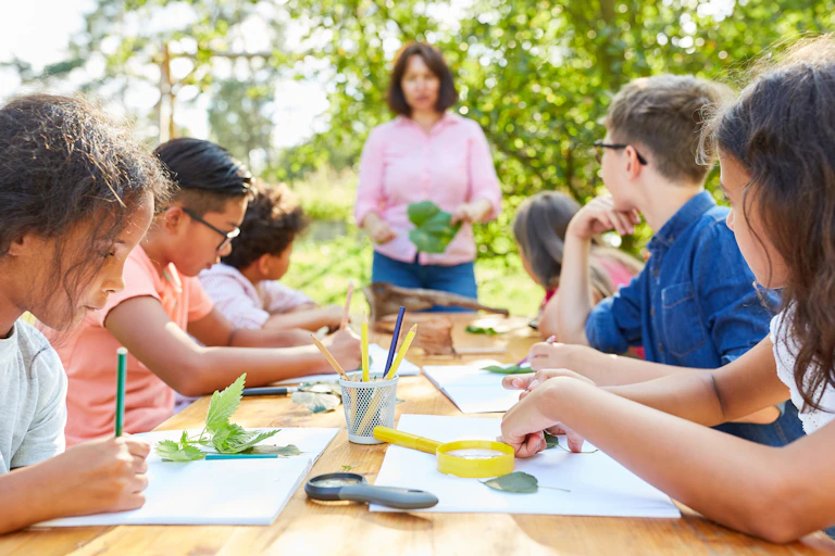 Six children sitting around table outside with paper, leaves, female teacher standing holding greenery at far end of table