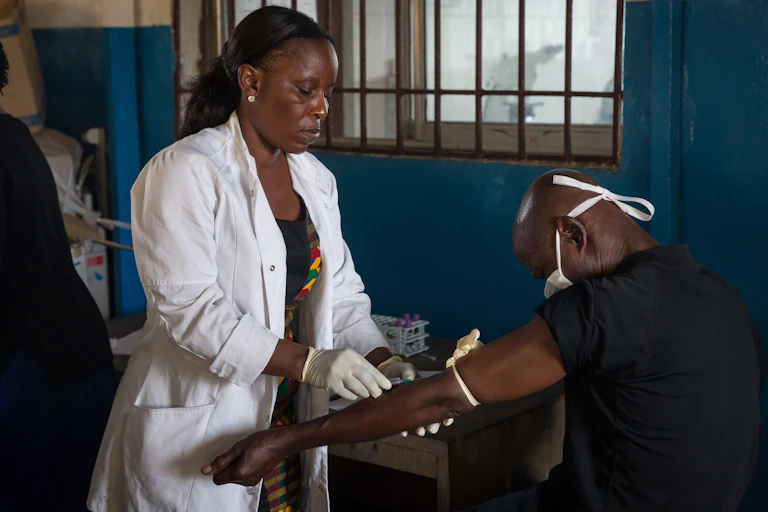 Woman in a white coat and gloves holds the arm of a man who is wearing a face mask