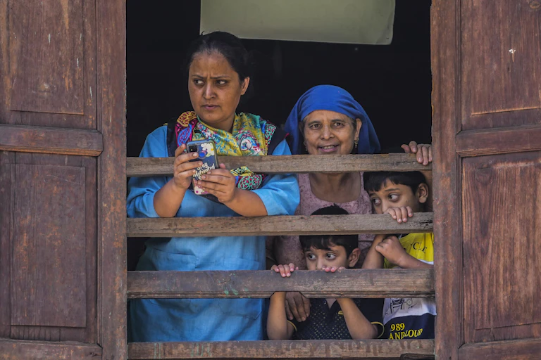 Two adults and two children look out a window of a house. A woman in a blue dress holding a smartphone ooks skeptical.