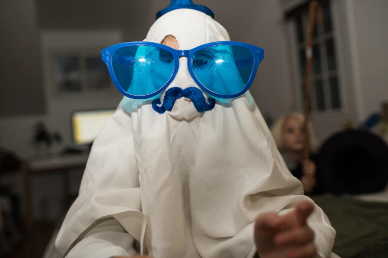 A child dressed as a ghost in a white sheet with giant blue sunglasses and a blue moustache.