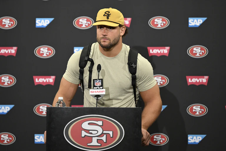 A muscular man in a pale green T-shirt wearing a ball cap stands behind a microphone.