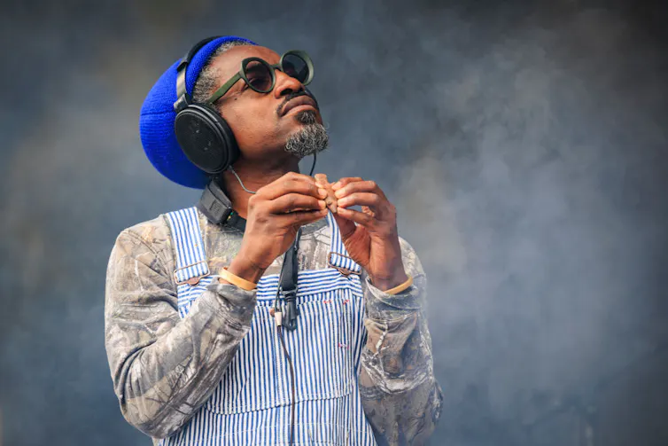 Un homme noir portant des lunettes de soleil, des écouteurs et un bonnet bleu saisit une petite flûte avec les doigts de ses deux mains