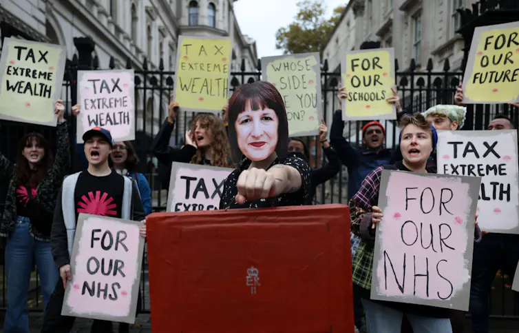Protestors hold placards, with one wearing a Rachel Reeves mask.