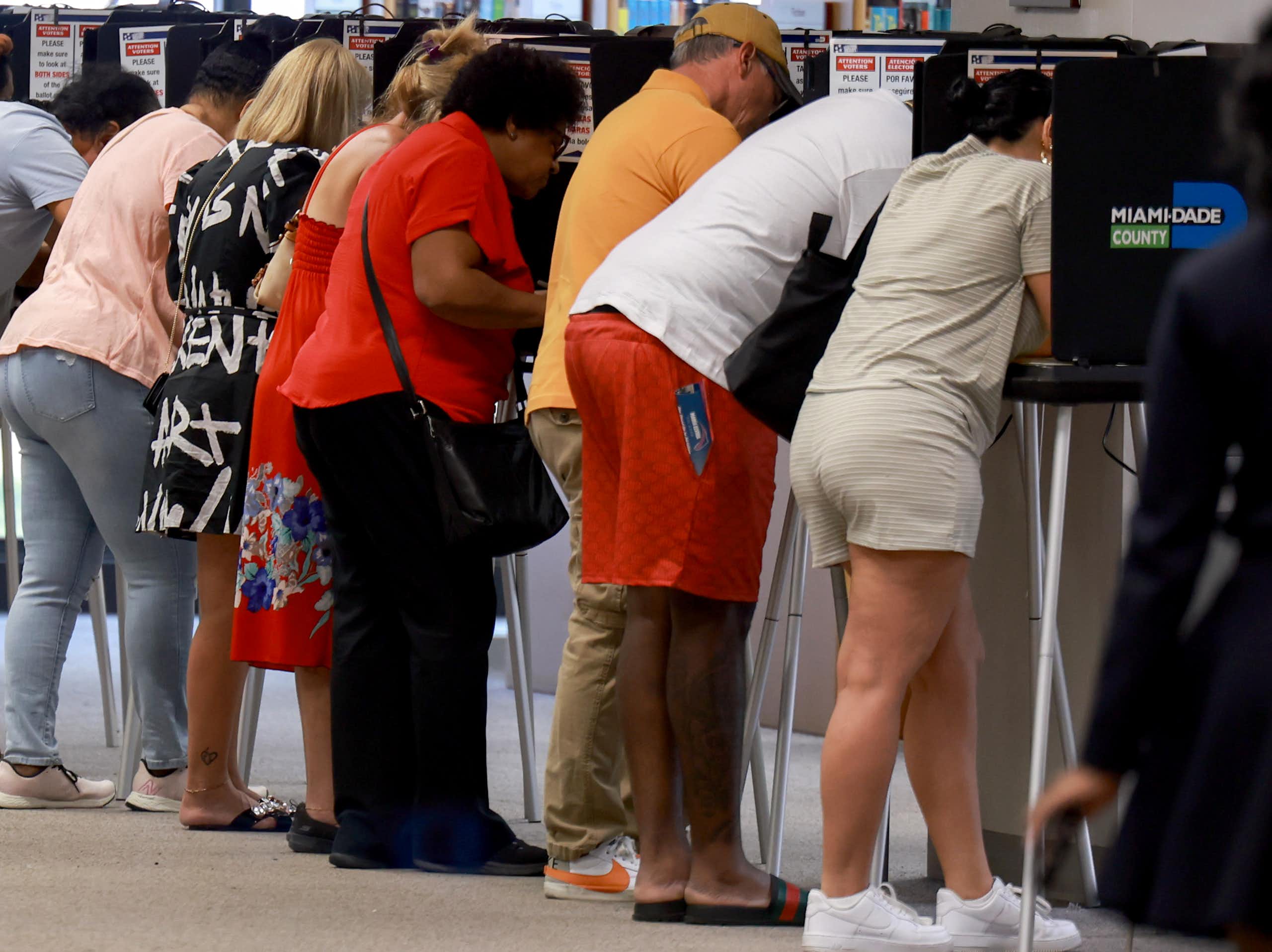 A row of people are seen facing away from the camera and leaning over little tables that are surrounded by black barriers.