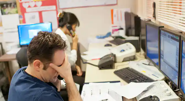 Man in blue scrubs looks stressed out while woman with hair in a bun speaks on the phone.