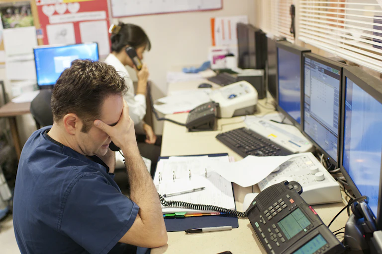 Man in blue scrubs looks stressed out while woman with hair in a bun speaks on the phone.