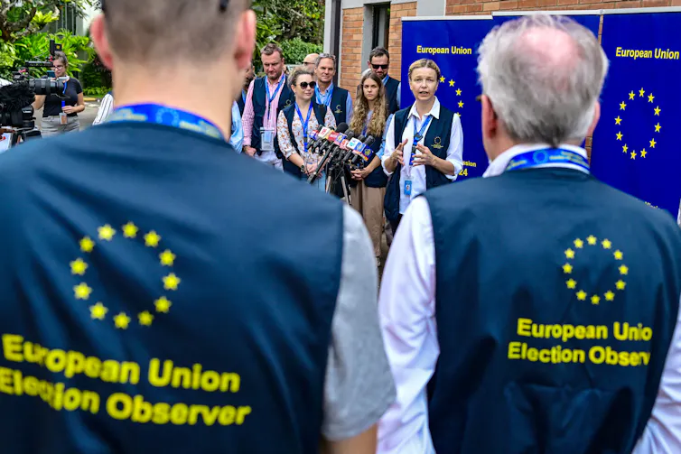 People wearing blue vests with yellow stars that say 'European Union Election Observer' look at a woman who is speaking to a crowd.