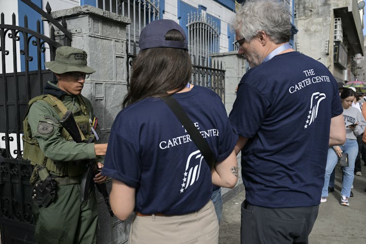 A man and a woman wear navy blue shirts that say 'Carter Center' in white and face a man wearing a camouflage green uniform outside of a large gate.