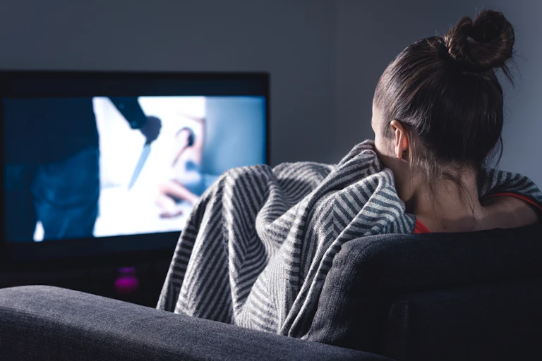 Mulher no sofá, enrolada em um cobertor, assistindo a um filme de terror na TV