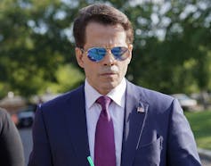 A dark-haired man with reflective aviator sunglasses in a blue suit and purple tie.