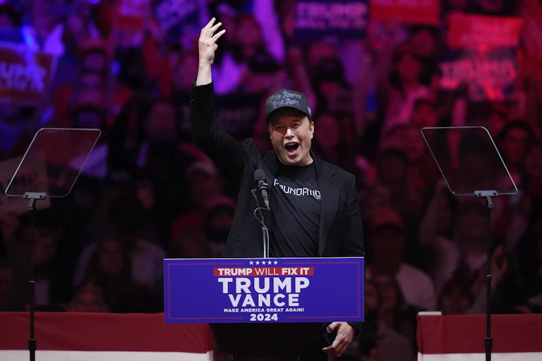 Man wearing black Make America Great Again hat and a black jacket, with his hand raised and mouth open at a lectern.