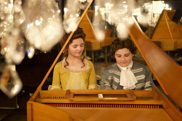 A young boy and girl play piano under a chandelier.