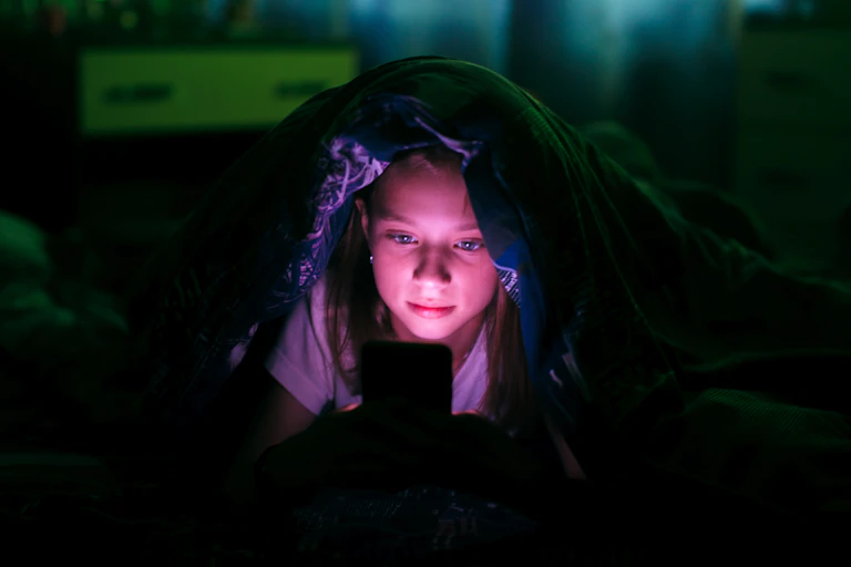 A teen girl with blanket over her head interacting with phone screen at night.