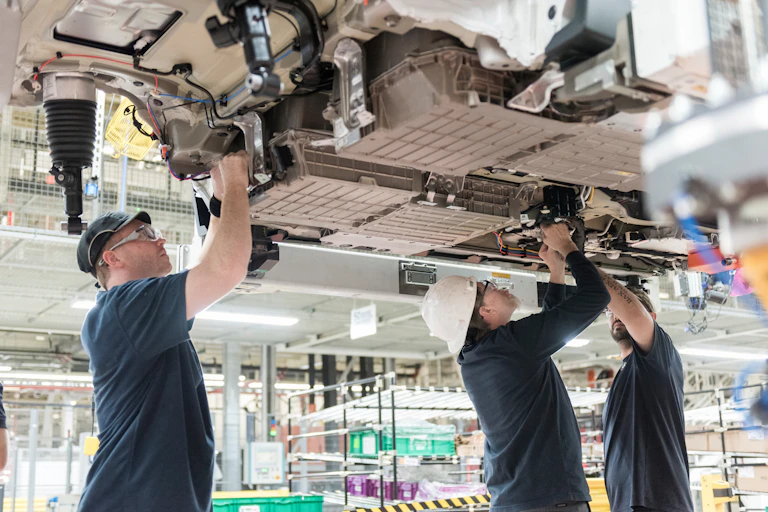 Three workers install a battery under a vehicle being built.