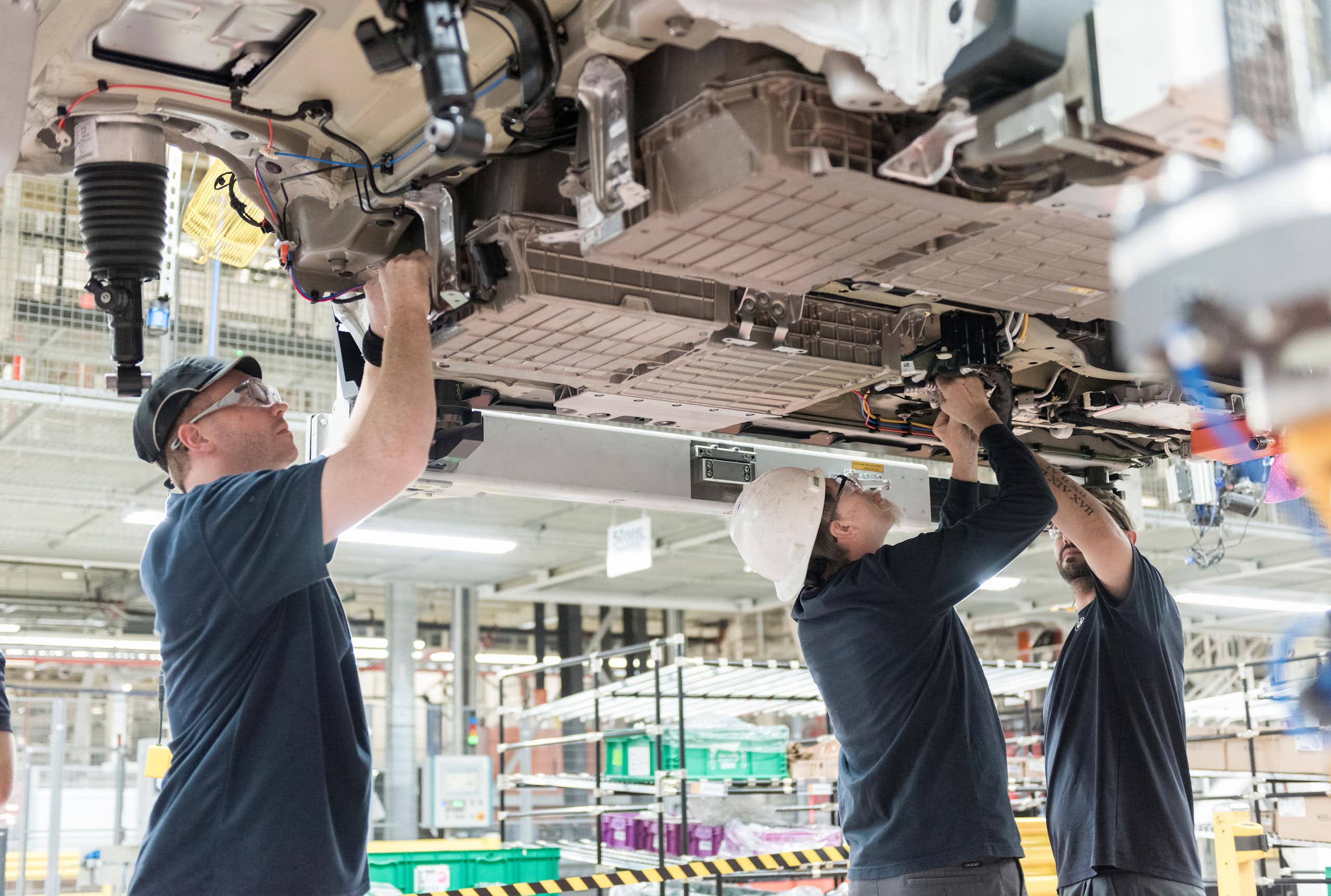 Three workers install a battery under a vehicle being built.