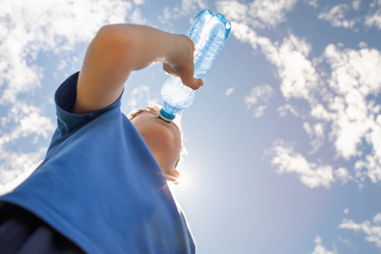 A boy drinks from a bottle of water against the blue sky.