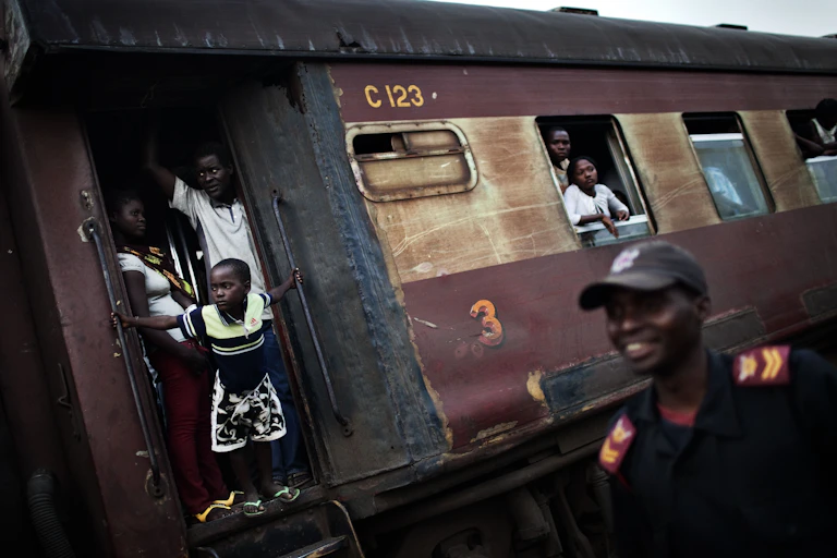 A child leans out of a train door and passengers stand in the windows, a security guard smiling in the foreground.