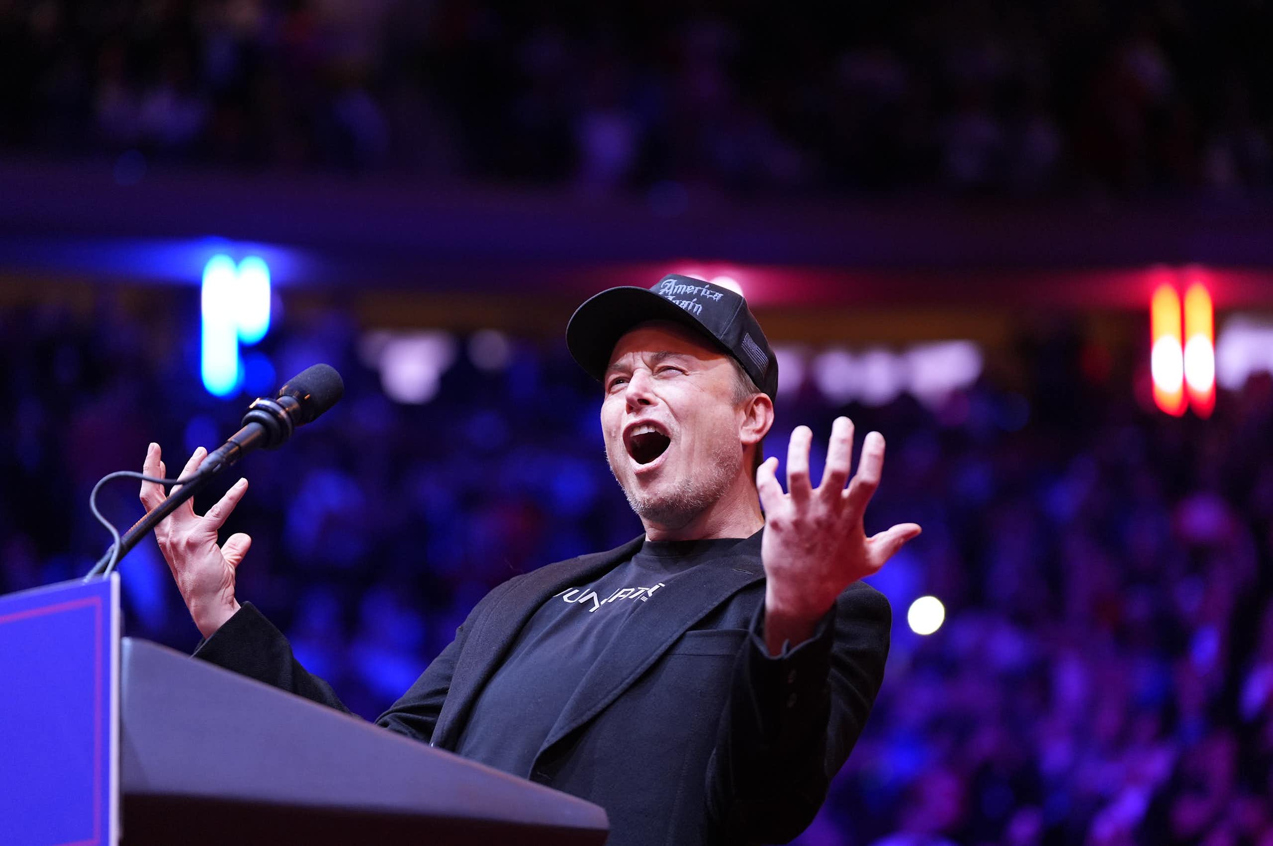 Man in black baseball hat, black T-shirt and black blazer speaks expressively at lectern