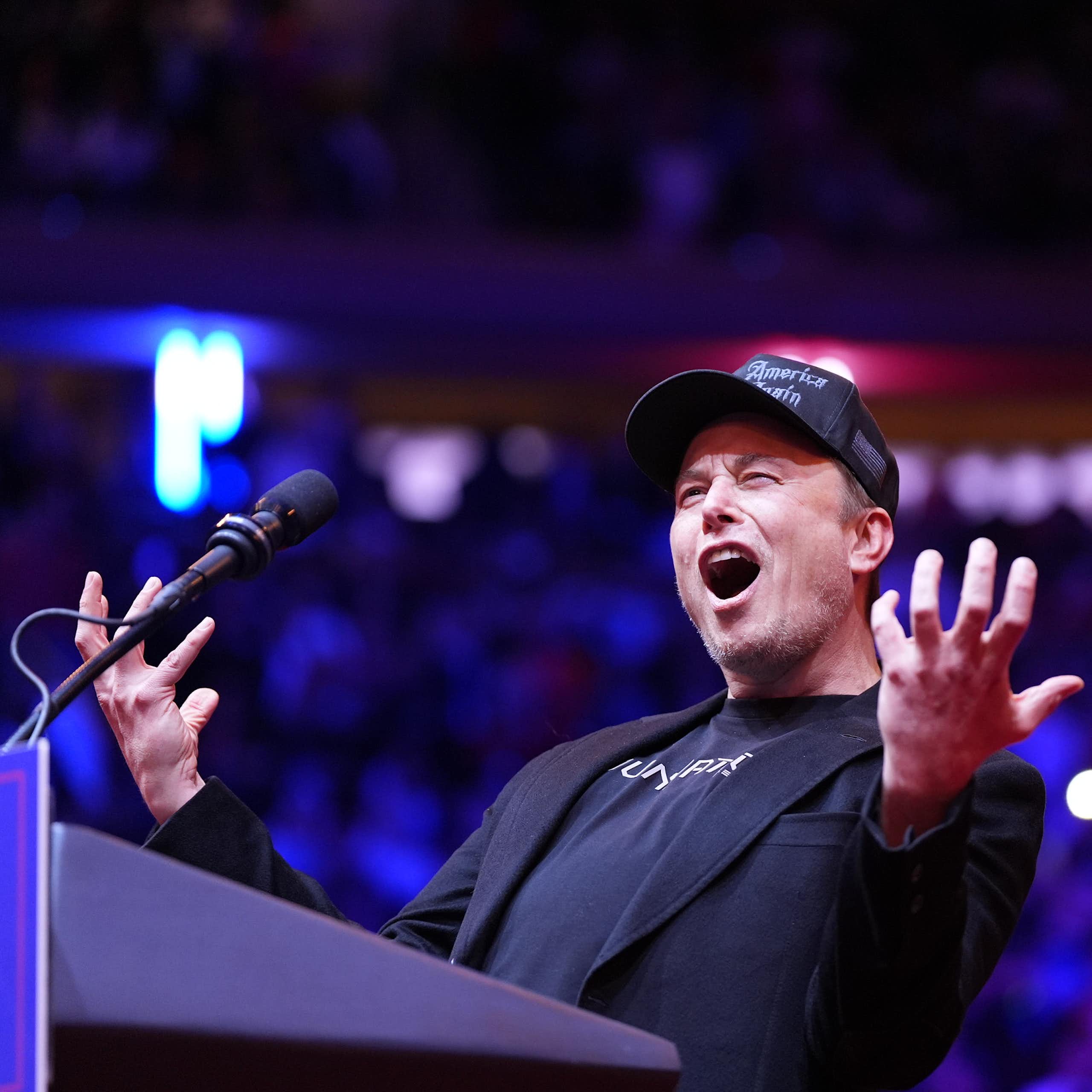 Man in black baseball hat, black T-shirt and black blazer speaks expressively at lectern